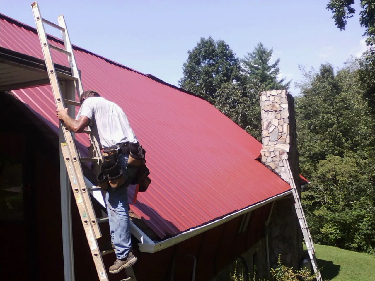 Roofer on a ladder beside a red metal roof and stone chimney — Soffit & Fascia Repair by Roofing Brothers in Berkley